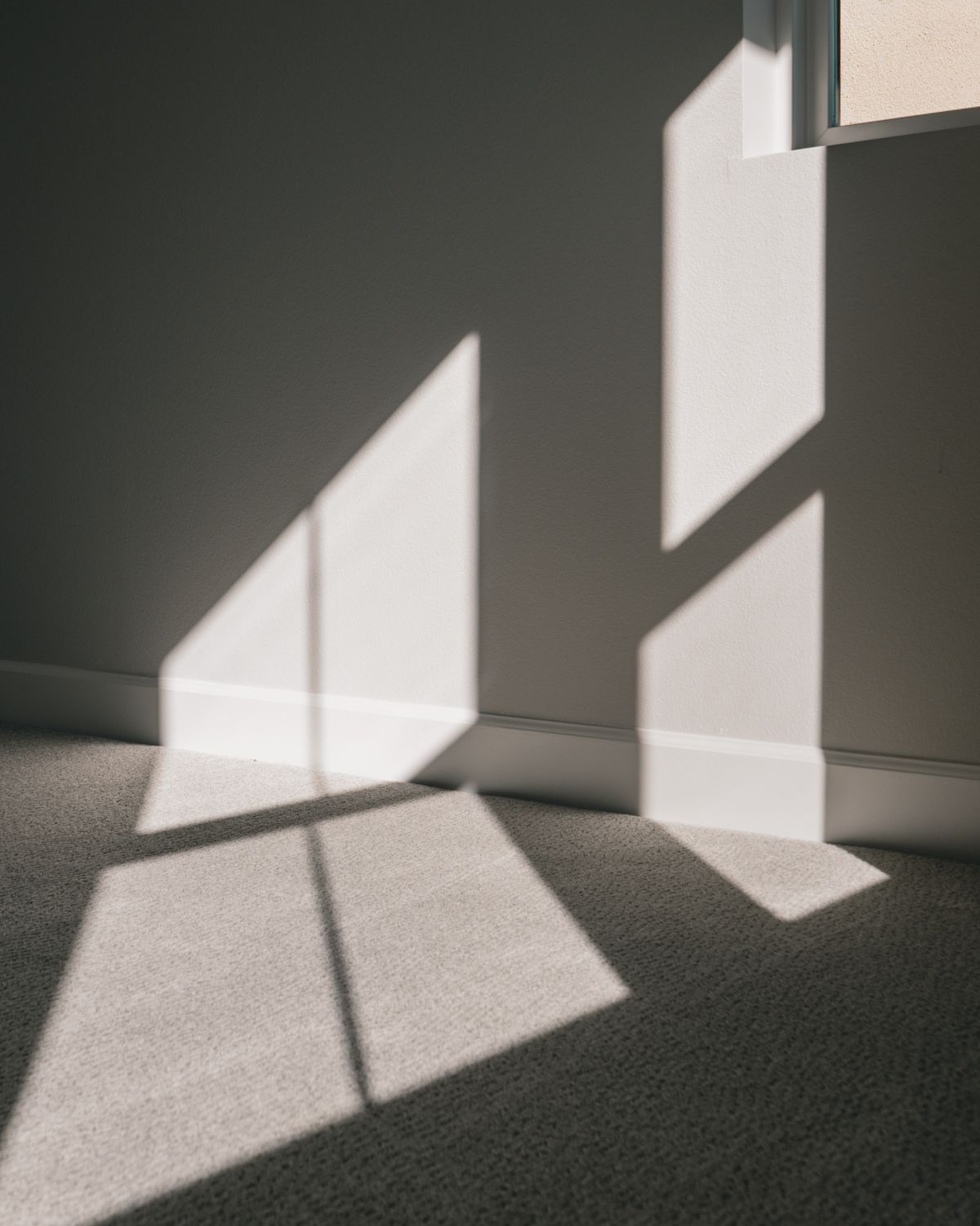 A hard-edged patch of direct sunlight thrown from a window across a gray carpet and up the wall beside it, the frame of the window printed on the floor as sharp bright panels